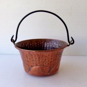 May include: A small, hammered copper bucket with a black metal handle. The bucket has a textured surface and a rounded base. The handle is arched and attached to the bucket with small metal loops. The copper has a warm, reddish-brown hue.
