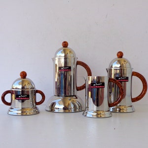 May include: A set of four stainless steel coffee pots and mugs. Each piece has a wooden handle and a wooden knob on top. The mugs and pots have a shiny, reflective surface. The text "orchidea" is visible on each piece.