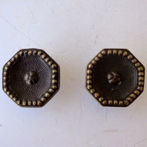 Könnte beinhalten: Zwei achteckige Schrankknöpfe aus antikem Bronze. Jeder Knopf hat eine strukturierte Mitte mit einem kleinen floralen Design und ist von einem Rand aus kleinen, erhabenen Punkten umgeben. Die Knöpfe sind vor einem weißen Hintergrund platziert.