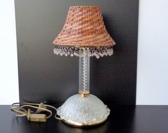 May include: A table lamp with a woven wicker lampshade and a clear glass base with crystal accents. The lamp has a gold-colored base and a clear cord.