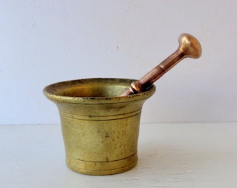 May include: A vintage brass mortar and pestle. The mortar is a small, cylindrical bowl with a flared rim and horizontal grooves. The pestle is a rod with a rounded head. The set has a warm, antique appearance.