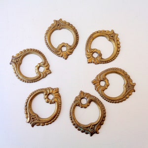 Może przedstawiać: Sześć antycznych uchwytów do szuflad w odcieniu mosiądzu ułożonych w okrąg. Każdy uchwyt ma ozdobny, ażurowy wzór z centralnym otworem i dekoracyjnymi detalami na krawędziach. Okucia są na białym tle.