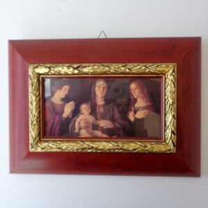 May include: A framed print of a Renaissance-style painting. The frame is a dark reddish-brown with a gold-colored inner border. The artwork depicts three figures, one holding a baby.