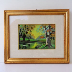 May include: A framed landscape painting with a gold-colored ornate frame. The artwork depicts a serene scene with a river, trees, and colorful foliage. The painting is set against a neutral-colored mat.