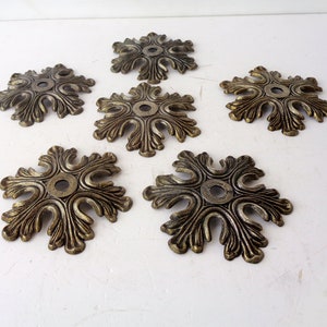 Może przedstawiać: Sześć antycznych, mosiężnych rozet dekoracyjnych z kwiatowym wzorem. Każda rozeta ma centralny otwór i ozdobne, zakrzywione detale w kształcie liści. Rozety ułożone są na białej powierzchni, prezentując swoje postarzone, metaliczne wykończenie.