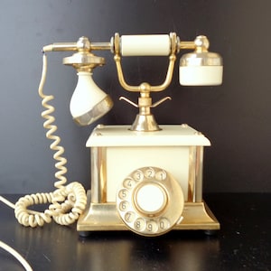 Può includere: Un telefono a disco vintage con base bianca e dettagli dorati. Il telefono ha una cornetta bianca e un disco bianco con numeri neri.