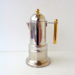 Puede incluir: Una cafetera italiana de acero inoxidable con asa y tapa doradas. La cafetera tiene una base redonda y una tapa abovedada con un pomo dorado. Diseñada para preparar espresso, es un electrodoméstico de cocina clásico.
