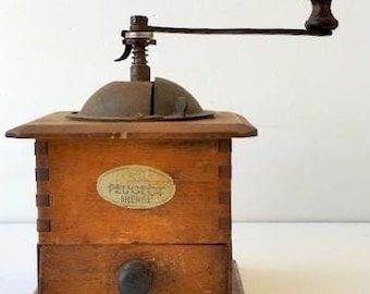 May include: Vintage wooden coffee grinder with a metal handle and top. The grinder has a small drawer and an oval label with text. The wood is a warm brown color, and the metal components show signs of age.