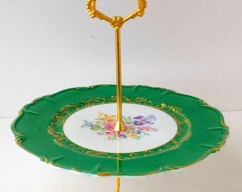 Puede incluir: Una bandeja de dos niveles con asa y soporte dorados. El nivel superior es un plato con borde festoneado en verde esmeralda con ribete dorado. El centro del plato presenta un diseño floral. La bandeja tiene aproximadamente 25 cm de diámetro.
