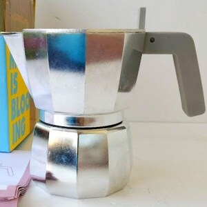 Op de afbeelding: Een zilveren espressomaker voor op het fornuis met een grijze handgreep en deksel. De koffiemaker is gemaakt van glanzend metaal en heeft een geometrisch ontwerp. Een blauwe doos met de tekst "IS BLOOMING" is zichtbaar op de achtergrond.