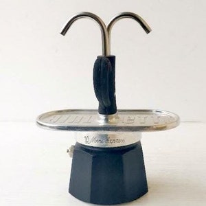 May include: A black and silver stovetop espresso maker. The coffee pot has a black base, a silver top, and a curved handle. The top handle is black. The espresso maker is designed to brew coffee on a stovetop.