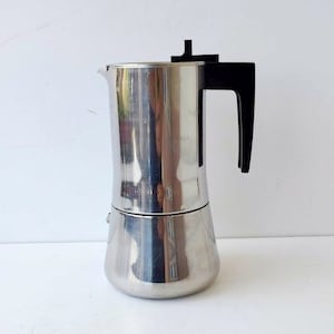 以下が含まれることがあります： 黒いハンドルと蓋が付いたステンレス製のエスプレッソメーカー。コーヒーポットは円筒形で、丸みを帯びたベースと注ぎ口が付いています。磨かれた金属は周囲の環境を反射し、光沢のある外観を作り出しています。