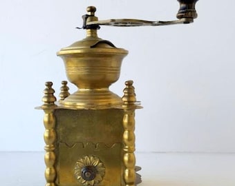 May include: Antique brass coffee grinder with a wooden handle. The grinder has a square base with decorative floral and column details. The top is a rounded shape with a crank arm. A vintage kitchen decor item.