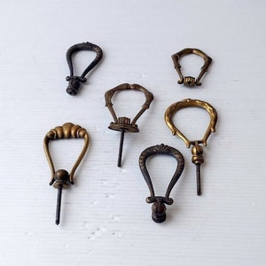May include: An assortment of six antique brass and bronze drawer pulls. The pulls feature various ornate designs, including a teardrop shape, and are attached to threaded screws. The finishes range from dark bronze to aged gold.