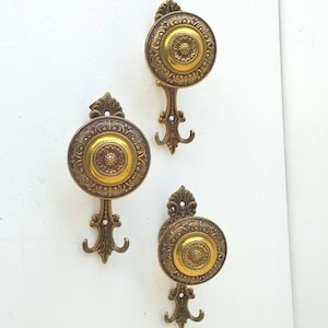 Puede incluir: Tres perchas de pared antiguas de latón. Cada percha presenta un diseño circular con un patrón floral central y detalles ornamentados. Las perchas están montadas sobre una superficie blanca, mostrando su estética vintage.