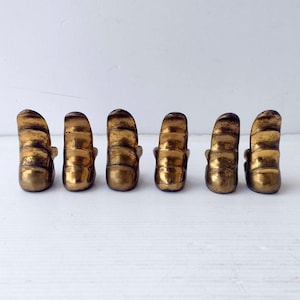 May include: Six antique brass drawer pulls. Each pull has a curved, ribbed design with a small, decorative element at the base. The pulls are arranged in a row against a white background, showcasing their aged, golden finish.