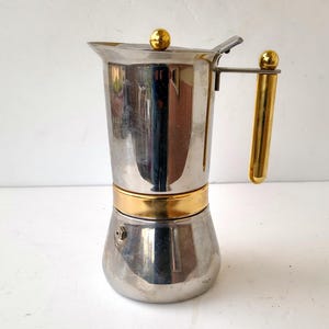 Pode incluir: Uma cafeteira italiana em aço inoxidável com detalhes dourados. A cafeteira tem um corpo cilíndrico com uma faixa dourada, uma alça dourada e um botão dourado na tampa. Projetada para preparar café expresso.