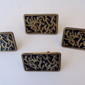 Puede incluir: Cuatro tiradores rectangulares para armarios con fondo oscuro y diseño floral dorado ornamentado. Los tiradores tienen forma ligeramente inclinada y estética vintage, adecuados para restauración de muebles o proyectos decorativos.