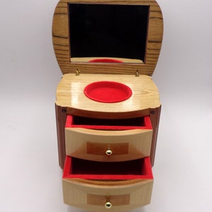 Könnte beinhalten: Hölzerne Schmuckschatulle mit Klappdeckel und Spiegel. Die Box hat zwei Schubladen mit rotem Samtfutter und goldfarbenen Knöpfen. Das Äußere weist eine Kombination aus hellen und dunklen Holztönen auf.