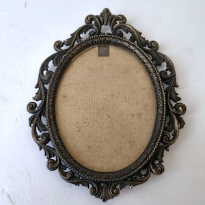 Puede incluir: Marco de fotos ovalado y ornamentado con acabado bronce oscuro. El marco presenta intrincados adornos y una superficie texturizada. El interior contiene un respaldo beige, listo para una fotografía o una obra de arte. Una pieza decorativa clásica.