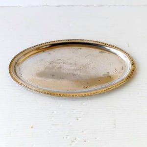 Può includere: Vassoio ovale argentato con bordo dorato bordato di perline. Il vassoio ha una superficie riflettente ed è posto su uno sfondo bianco. Il vassoio è probabilmente vintage o antico.