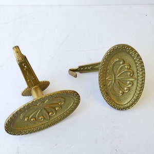 Puede incluir: Dos abrazaderas de cortina antiguas de latón. Cada abrazadera presenta una placa decorativa ovalada con un diseño floral en relieve y un vástago. Las abrazaderas son de color dorado cálido sobre un fondo blanco.