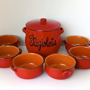 Può includere: Un set Fagiolata in ceramica rossa, composto da una pentola con coperchio e sei ciotole. La pentola ha la scritta "Fagiolata" in nero. Le ciotole e la pentola hanno interni arancioni e piccole maniglie.