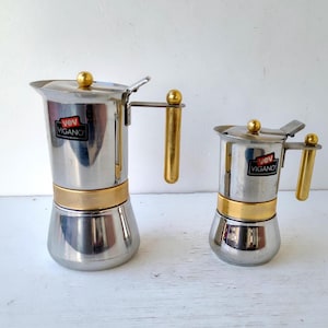 Pode incluir: Duas cafeteiras de aço inoxidável da Vev Vigano. Cada cafeteira apresenta um acabamento prateado polido, uma faixa central dourada, uma alça cilíndrica dourada e um botão dourado na tampa articulada. Uma etiqueta com o logotipo preto e vermelho da Vev Vigano está afixada na frente de cada unidade.