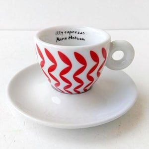 Puede incluir: Juego de taza y platillo de espresso blancos. La taza presenta un patrón de zigzag rojo y el texto "illy espresso Mono Hotoun". El asa tiene un diseño circular.