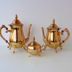 May include: A gold-colored tea set, including a teapot, a coffee pot, and a sugar bowl. The set has ornate handles and decorative feet. The lids are topped with a decorative finial. The set is made of a shiny, reflective material.