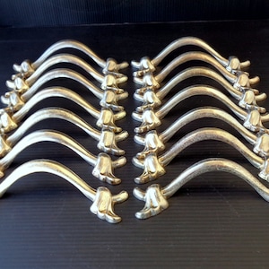 Könnte beinhalten: Eine Ansammlung von zwölf goldfarbenen Schrankgriffen, angeordnet in zwei Reihen. Jeder Griff hat ein geschwungenes Design mit einem dekorativen floralen Akzent an jedem Ende. Die Griffe sind aus Metall und werden auf einer dunklen Oberfläche präsentiert.