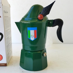 May include: A green stovetop espresso maker with a black handle and a decorative leaf and berry accent. The espresso maker has a shield with the Italian flag colors. A box is partially visible in the background.