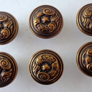 May include: Six antique bronze-colored cabinet knobs. Each round knob features a detailed, raised design of a central floral element surrounded by swirling patterns. The knobs have a vintage aesthetic.