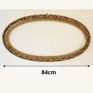 Peut inclure: Un cadre photo ovale de style antique avec des détails dorés ornés et tourbillonnants. Le cadre mesure 84 cm de long et est conçu pour contenir un portrait ou une œuvre d'art. Le cadre est suspendu sur un fond blanc.
