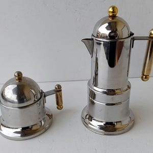 Puede incluir: Dos cafeteras italianas de acero inoxidable. La más grande tiene un asa y una tapa doradas. La más pequeña tiene un diseño similar. Ambas están sobre una superficie blanca.
