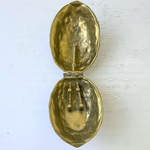May include: Open brass-coloured jewellery box in the shape of a walnut. The box is hinged and has a textured surface. The interior has two small posts for holding jewellery.