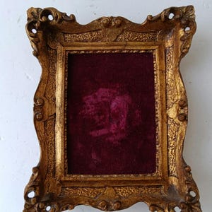 May include: An antique-style gold picture frame with a burgundy velvet backing. The frame features an elaborate, vintage design with curved edges and decorative detailing. Ideal for displaying artwork or photographs.