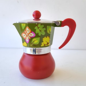 May include: A red and green stovetop espresso maker with a butterfly and leaf design. The top is green with a silver rim and a red handle and knob. The base is red.