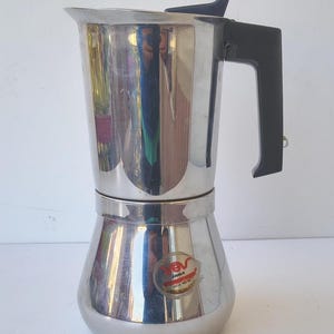 May include: A stainless steel stovetop espresso maker with a black handle and lid. The coffee pot has a classic design with a wide base and a narrow top. A small sticker with text is visible on the lower part of the pot.