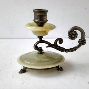 May include: An antique candle holder with a green and cream-colored base and accents. The candle holder has a decorative, curved handle with a floral design. The candle holder is made of metal and has a vintage aesthetic.