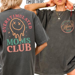 May include: Pepper-colored Comfort Colors t-shirt with the text "OVERSTIMULATED" arched above a melting smiley face and "MOMS CLUB" below. The front of the shirt features a smaller design with "OVERSTIMULATED" and "Mom's Club" text.