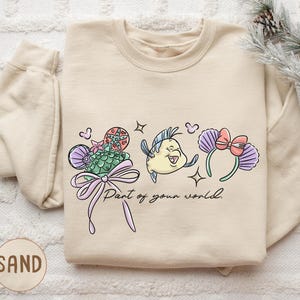 A beige sweatshirt with a cartoon fish design and the text "Part of your world" printed on it, along with a bow and other decorative elements.