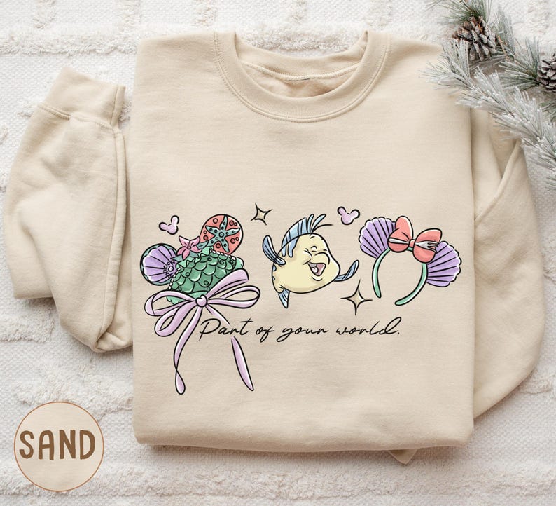 A beige sweatshirt with a cartoon fish design and the text "Part of your world" printed on it, along with a bow and other decorative elements.