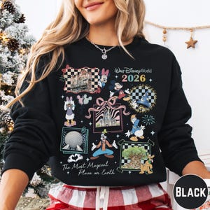 A woman wearing a black sweatshirt with a colorful graphic design featuring various Disney characters and landmarks, including a castle, a Mickey Mouse, and a fireworks display.