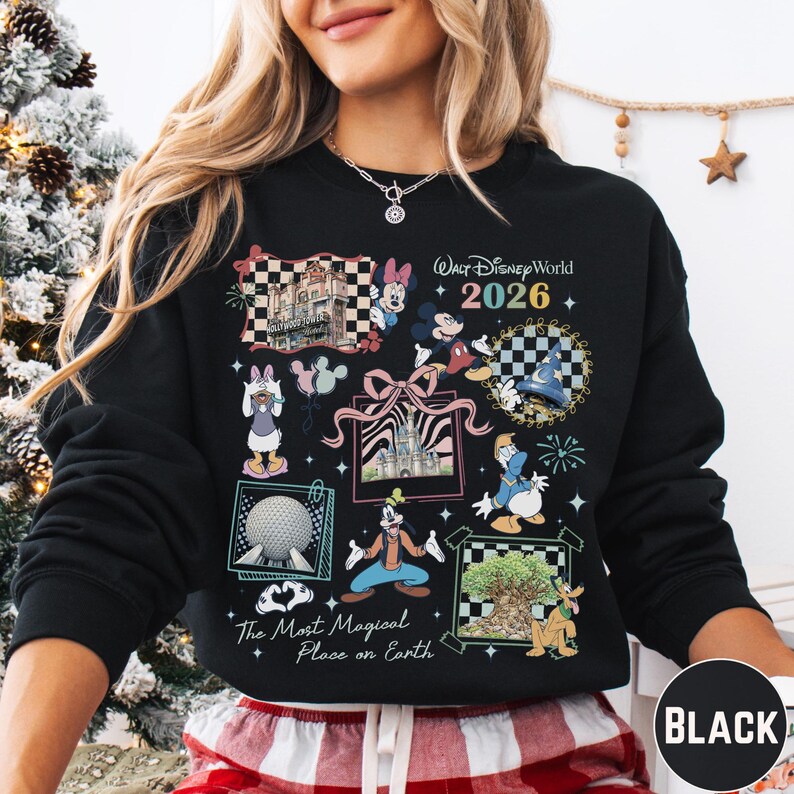 A woman wearing a black sweatshirt with a colorful graphic design featuring various Disney characters and landmarks, including a castle, a Mickey Mouse, and a fireworks display.