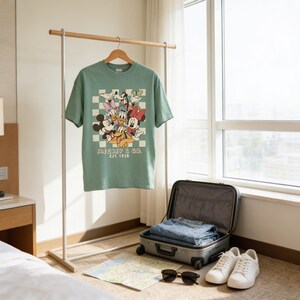 a green t-shirt hanging on a wooden hanger, with a pair of jeans and a pair of white sneakers placed next to it. The scene also includes a suitcase and a map on the floor.