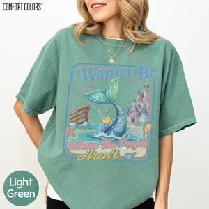 A woman wearing a green t-shirt with a graphic design featuring a mermaid, a castle, and the text "I Wanna Be Where The People Aren't".
