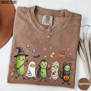 a brown t-shirt with a Halloween-themed design, including a witch, pumpkin, ghost, mummy, and a cat.