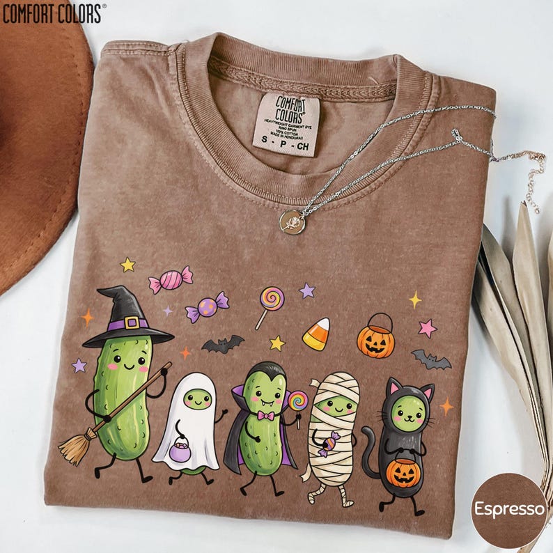 a brown t-shirt with a Halloween-themed design, including a witch, pumpkin, ghost, mummy, and a cat.