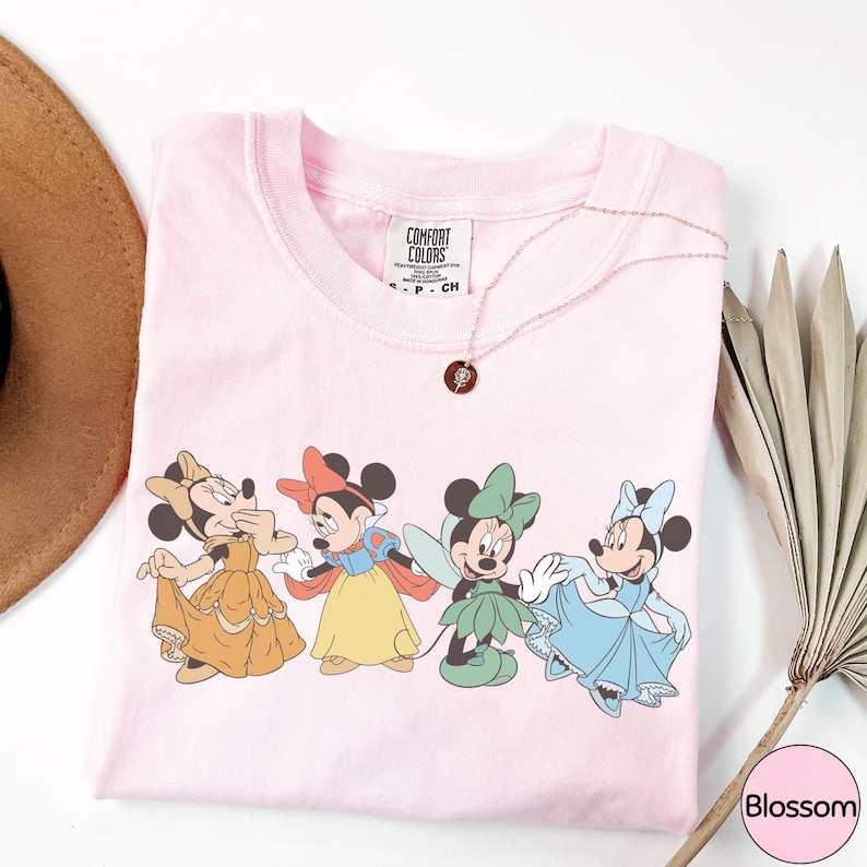 A pink t-shirt with a graphic of four Disney Minnie Mouse characters in different outfits, along with a necklace and a hat.